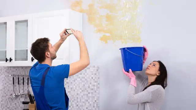 Homeowner photographing a water-stained wall with a phone while a person holds a bucket under a ceiling leak
