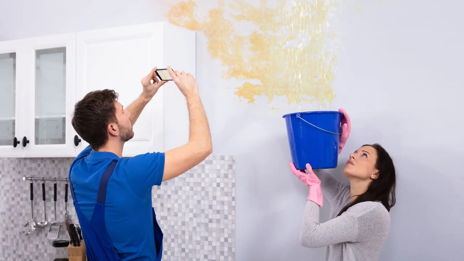 Homeowner photographing a water-stained wall with a phone while a person holds a bucket under a ceiling leak
