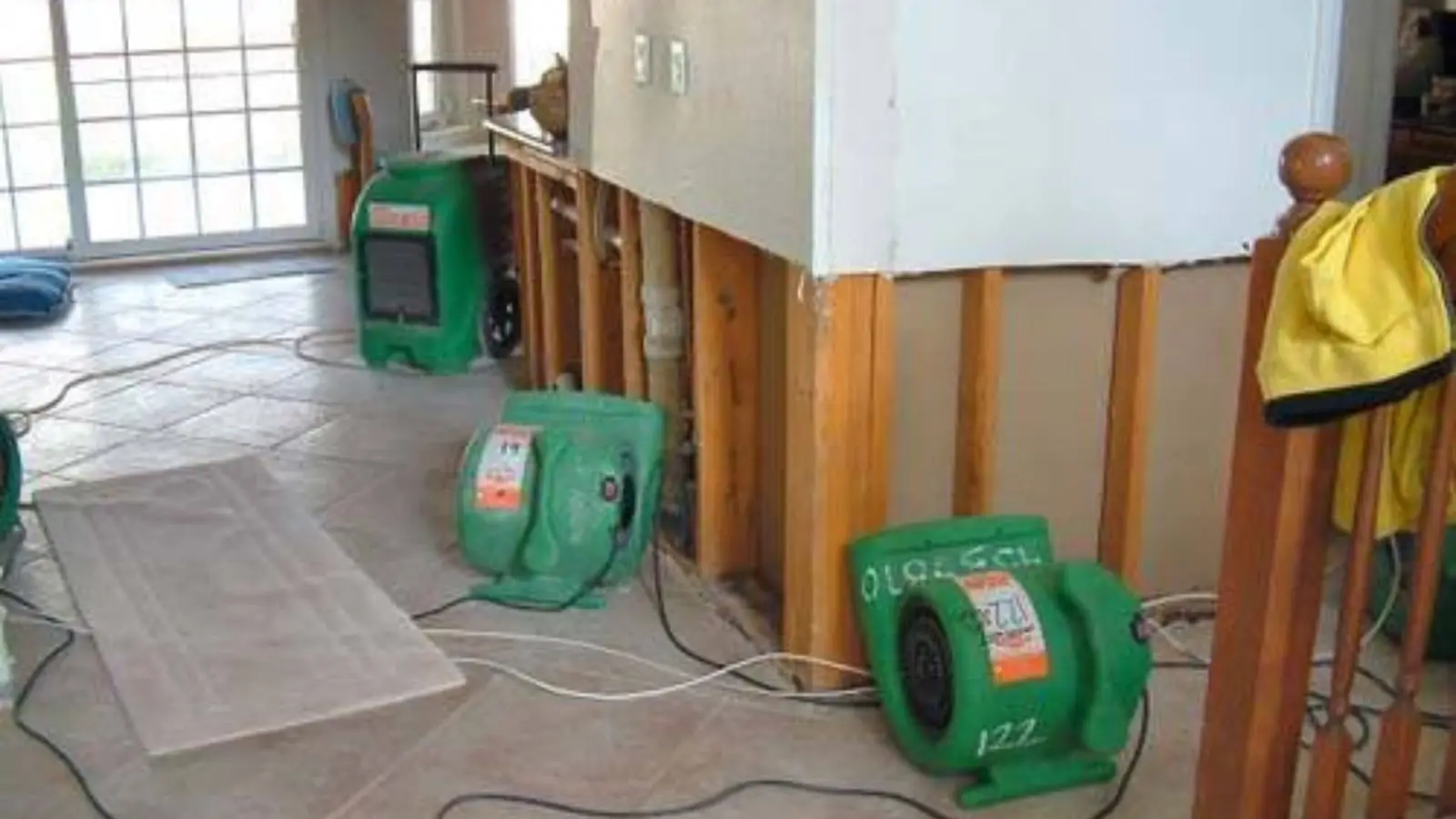 Green air movers drying a room with wet drywall cut away below the baseboard